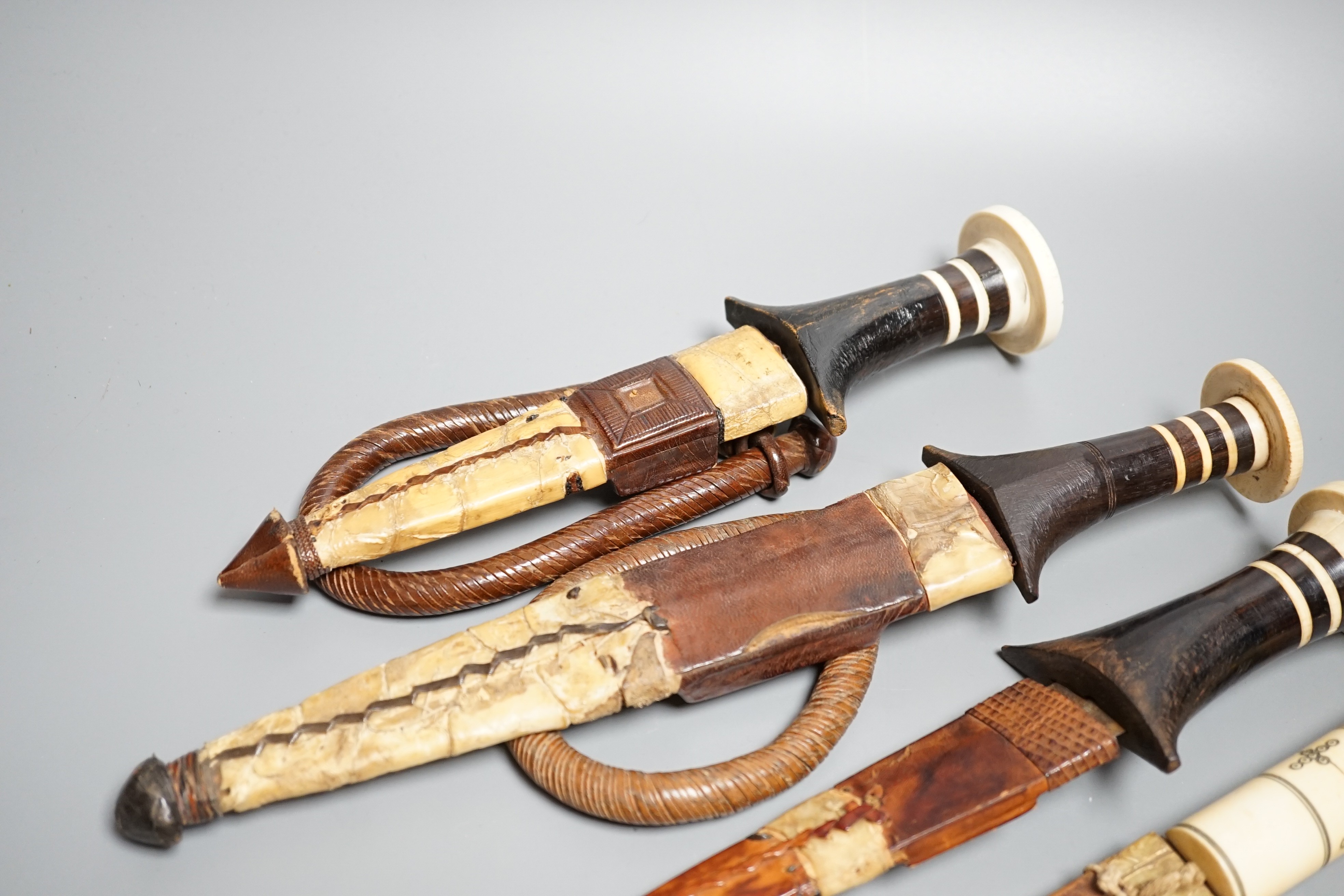 Five assorted African Nilotic tribal daggers c.1900, comprising three with turned ivory mounted ebony handles (two with silver pommels); and two further with cylindrical ivory handles, all in their leather sheaths, maxim